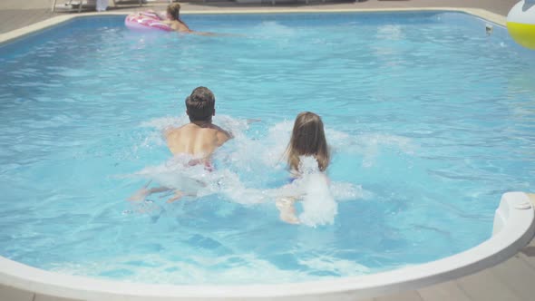 Back View of Young Caucasian Man and Woman Swimming in Outdoor Water Pool at Luxurious Resort