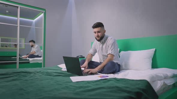 Wide Shot of Busy Young Handsome Businessman Analyzing Documents and Emarket Sitting on Bed in Green