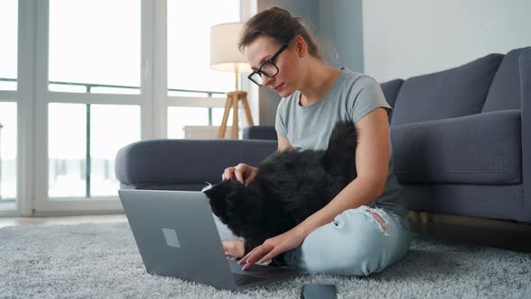 Casually Dressed Woman with Glasses Sits on a Carpet with a Laptop and Strokes a Fluffy Black Cat