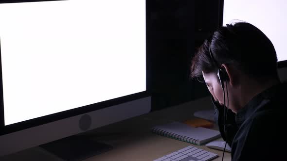 Asian man in headset working at late night in dark office, being tired and sleepy, rubbing eyes