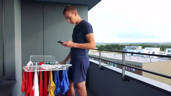 A Young Man Takes Down Clothes From the Drying Rack on the Balcony and Works on the Smartphone