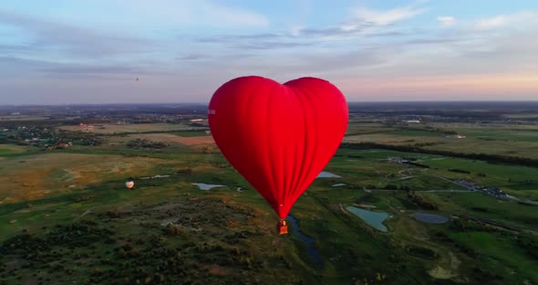 Aerostat in the hear form in the air. Red balloon fly over green meadows with lakes at sunset. Aeria