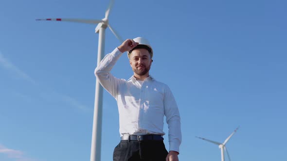 Air Power Engineer Stands in Business Style Clothes and White Helmet on a Background of Windmill
