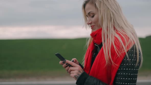 Blonde Woman Is Walking on the Road and Using the Smartphone. Girl with a Red Scarf Look at the
