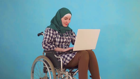 Young Muslim Woman Disabled in a Wheelchair Uses a Laptop on a Blue Background