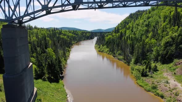 A drone flies underneath a bridge and high above the water of a river surrounded by trees and mounta