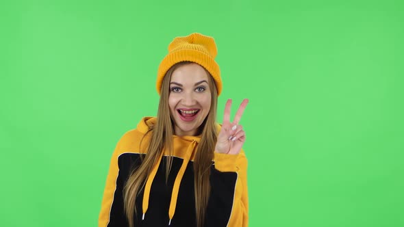 Portrait of Modern Girl in Yellow Hat Is Showing Two Fingers Victory Gesture. Green Screen