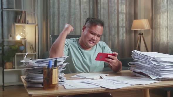 Fat Asian Man Being Upset Losing Game On Smartphone After Working With Documents At The Office