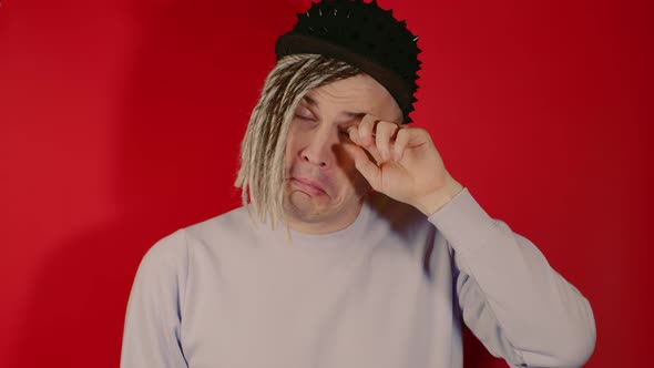 Young Man in Cap Rubs His Eye Looking at Camera on Red Background in Studio