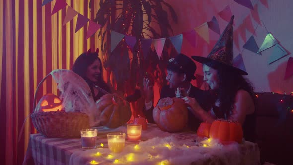 Two Girls and a Guy in Creepy Halloween Costumes are Sitting at a Table Chatting and Carving Faces