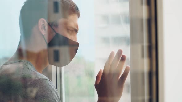 Young Caucasian guy in a medical mask is fit to a closed window during the quarantine and isolation