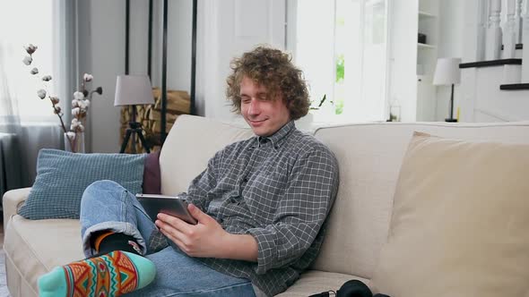 Man Dressed in Casual Clohes Sitting on the Cozy Sofa in Contemporary Apartment and Using Tablet PC