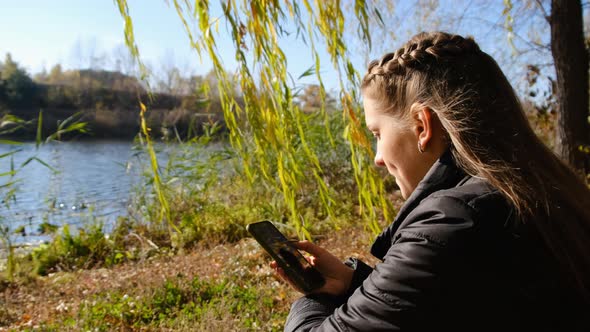 Young Woman with a Smartphone in Hands Sits on Yellow Foliage By River in Autumn