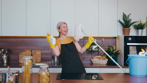 Cheerful Grayhaired Asian Housewife Wearing an Apron and Gloves Dancing in Kitchen with a Mop and