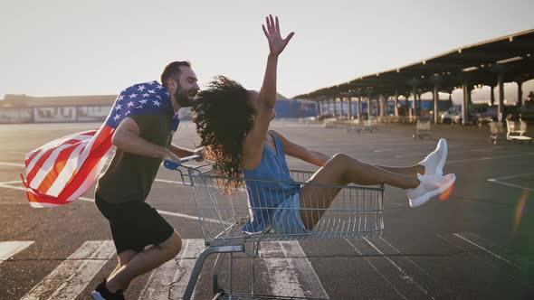 Young Guy Wrapped in Flag of USA is Racing on Shopping Trolley with Darkskinned Girlfriend in It at