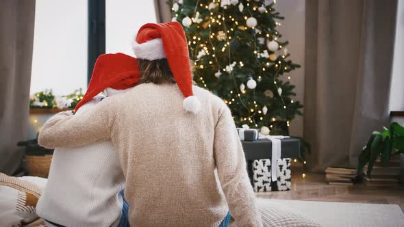 Young Unknown Man and Woman in Santa Hats are Hugging While Sitting at Home Against Green Decorated