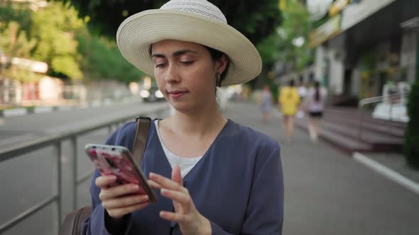 Portrait of a young woman in a straw hat standing on the street and swiping on a smartphone.