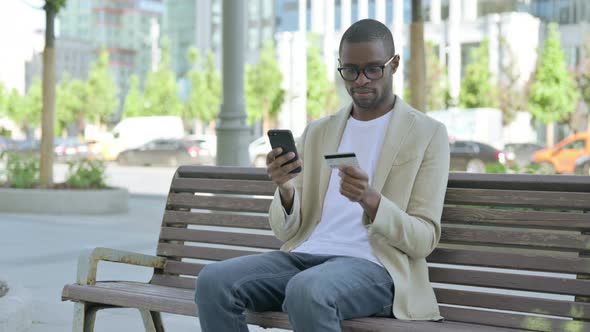 Excited African Man Shopping Online Via Smartphone While Sitting Outdoor on Bench