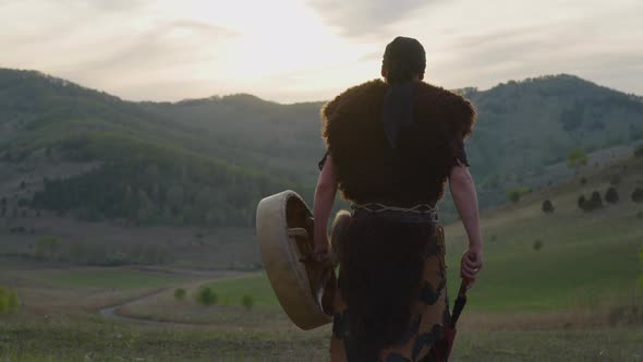 Actor Walks Carrying Shamanic Drum Against Old Hills