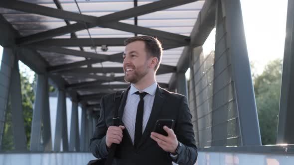 A Young Man in Business Clothes Walks Across the Bridge and Smiles