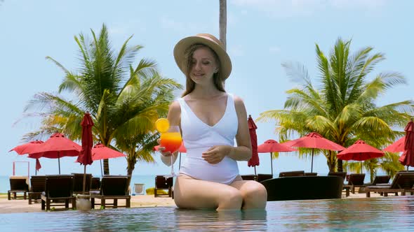 Young Woman in White Swimsuit Drinks Red Orange Nonalcoholic Cocktail Relax Sitting in Swimming Pool