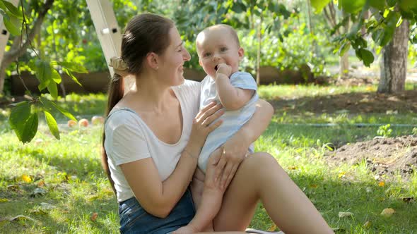 Portrait of Happy Smiling Mother Holding and Kissing Her Baby Son While Relaxing Under Apple Tree in