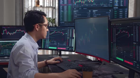 Asian Young Male Stock Market Broker Analyzing Graphs On Multiple Computer Screens