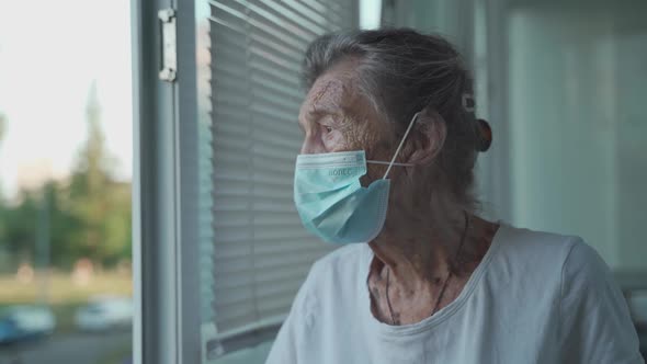 Sad Senior Woman 90 Years Old Wearing Protective Medical Mask Looking Out an Open Window in a