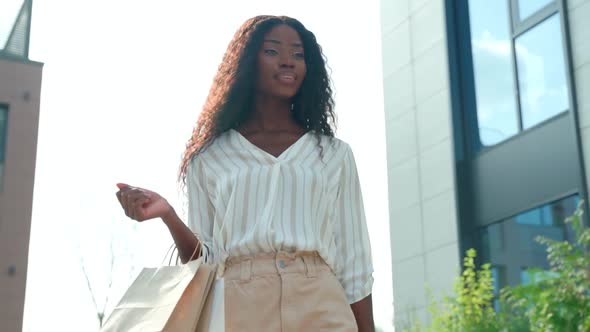 Happy Black Woman Shopper Walking on Street Holding Shopping Bags and Cellphone