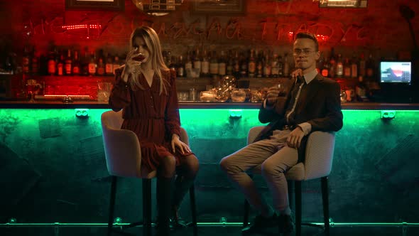 A Couple Having a Date in the Bar - Sitting on the Bar Chairs Towards the Camera and Drinking Their