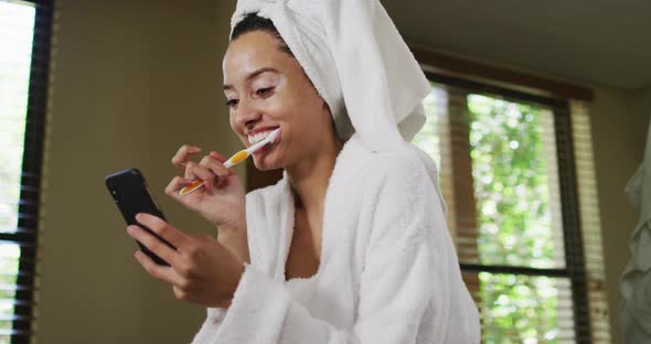 Happy biracial woman in robe brushing teeth and using smartphone