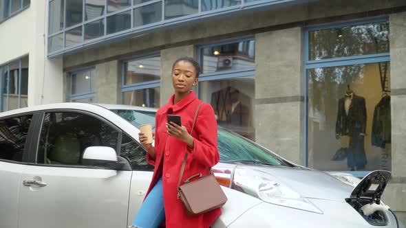 African American Girl Using Smartphone and Leans on Electric Car That Is Charging. Environmentally