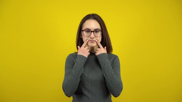 A Young Woman Makes a Sad and Cheerful Smile on Her Face with Her Fingers on a Yellow Background