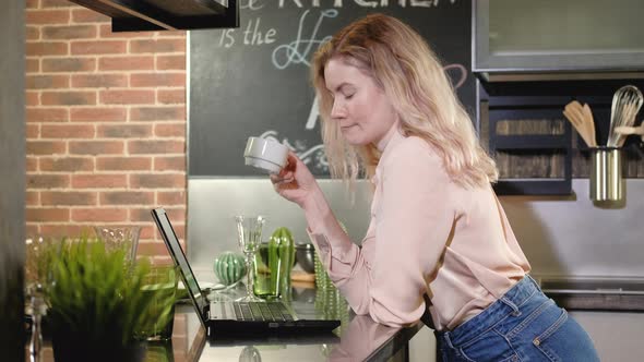 Woman drinks coffee in the kitchen in the evening and works with a laptop