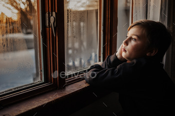  quarantined again due to the continuing pandemic. Boy at home looking out window at sunset.