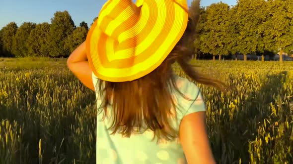 Beauty Girl Running in a Yellow Hat on Green Wheat Field Over Sunset Sky