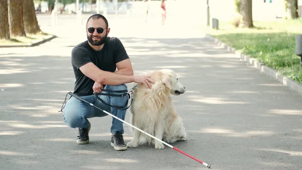 Blind Bearded Man with a Cane and Labrador Sitting on Pavement in Park