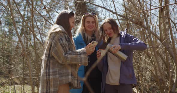 three girls forest spring drinking tea from thermos communicate smiling and looking phone