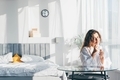 Pretty positive woman with long hair drinks water and sitting at glass table in spacious bedroom  - PhotoDune Item for Sale