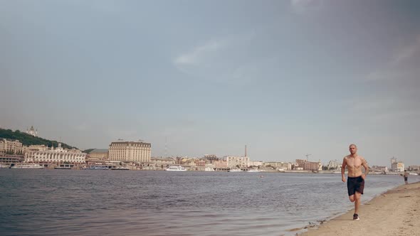 Muscular Young Man Running on the Beach in the City on a Sunny Day