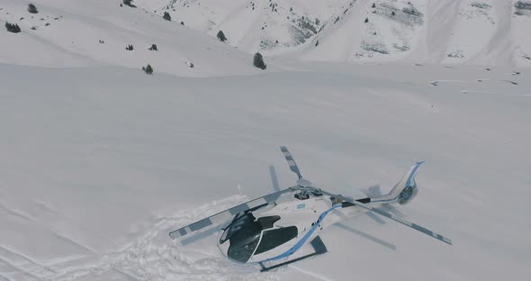 The aerial flies over a parked helicopter standing in the snow in the winter mountains