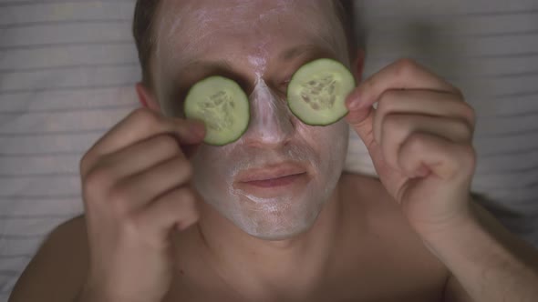 Young attractive man looks after his face, makes a cosmetic mask and puts a cucumber on his eyelids