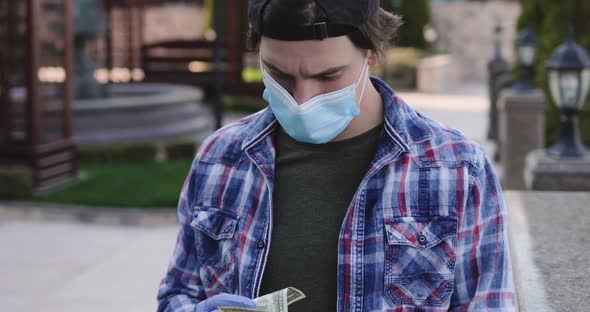 Man in Mask and Gloves Holds Last Dollar in Hands is Sad and Looking at Camera