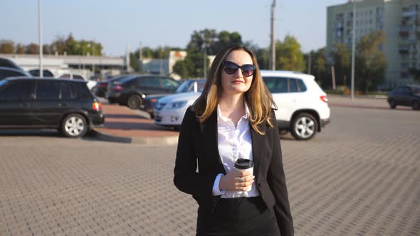 Young Smiling Businesswoman with Cup of Coffee Walking Near Auto Parking at Sunny Day. Happy