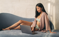 Asian woman sitting on couch in dressing gown, working on laptop at home against textured wall - PhotoDune Item for Sale