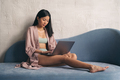 Asian woman sitting on couch in dressing gown, working on laptop at home against textured wall - PhotoDune Item for Sale
