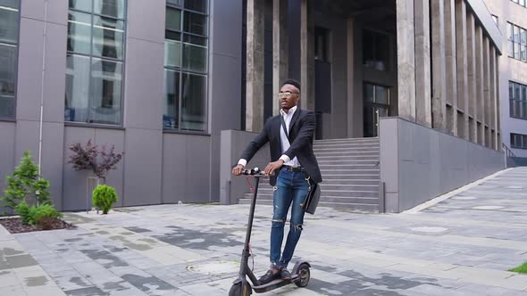 African American in Stylish Wear Riding on E-Scooter on the Urban Street Near Contemporary Building