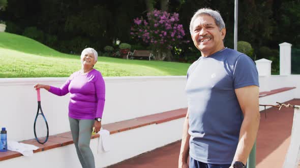 Video of happy biracial senior couple looking at camera on tennis court