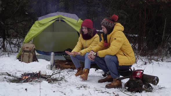 Young Man and Woman Warm Their Hands Over a Fire in the Winter Forest