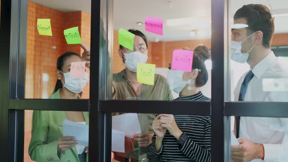 Group brainstorming of Asian people wearing mask, putting sticky note, put it on window in office.